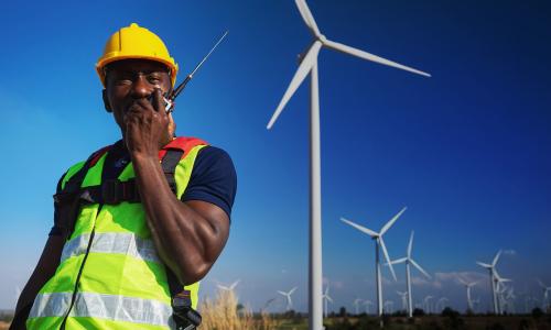 A person in a vest and hard hat in front of a wind turbine