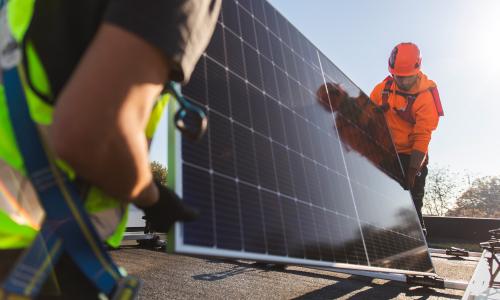 Two workers carrying a solar panel across a rooftop.