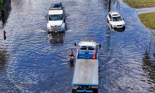 A person wades through water on a flooded street, next to partially submerged cars.